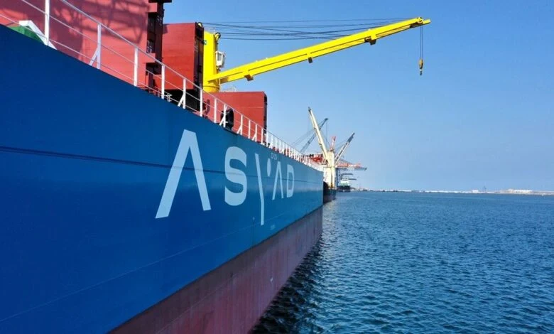 The image is a genuine news photo showing an Asyad-branded cargo vessel equipped with deck cranes, directly matching the article regarding Asyad Group and Asyad Shipping.