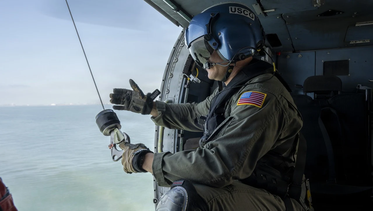 Clear news photo showing a US Coast Guard crew member in a blue USCG helmet and flight suit inside an aircraft during maritime operations over water, relevant to the article about USCG search and rescue technology.